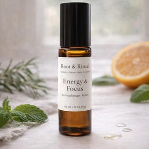 May include: Aromatherapy roller bottle with a black cap and a brown glass body. The label reads "Root & Ritual Energy & Focus Aromatherapy Roller 10 ml / 0.33 fl oz." The background includes a lemon slice and green herbs.