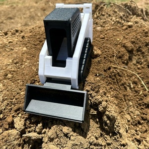 May include: A small, white and black toy skid steer loader with a black bucket and cab. The toy is sitting on a pile of dirt and is designed to resemble a real construction vehicle.