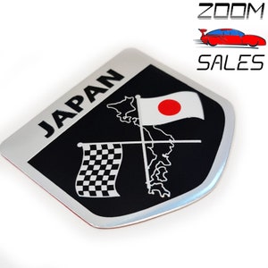 May include: A silver and black car emblem with the word "JAPAN" in white lettering. The emblem features a chequered flag, a map of Japan, and a Japanese flag with a red circle.