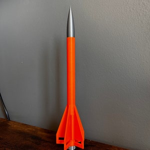 May include: An orange and silver model rocket with three fins. The rocket is standing upright on a wooden surface.