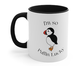 May include: White ceramic coffee mug with a black rim and handle. The mug has a black and white illustration of a puffin and the text "I'm so puffin lucky".