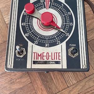 May include: Vintage Time-O-Lite Professional timer with a black and gold face. The timer features a red dial and a needle indicating the seconds. It has a focus switch and a push button. The timer is on a wooden surface.