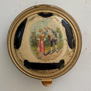 May include: A round, antique compact with a gold-tone metal frame. The lid features a painted scene of two figures in a garden, framed by black accents. The compact has a small clasp for opening.
