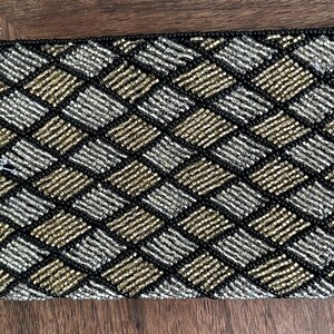 May include: Rectangular beaded clutch with a black zipper. The clutch features a woven pattern of silver, gold, and black beads. The beads are tightly arranged in a geometric design, creating a textured surface.