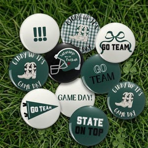 May include: A collection of round buttons with various designs and text, including "Go Team," "Game Day," and "State on Top." The buttons feature a green and white color scheme, with images of a cowboy boot, football helmet, and exclamation points. The buttons are arranged on a bed of green grass.