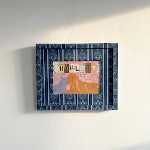 May include: A framed print with the name "CHARLOTTE" in block letters. The print features a dog and a cat illustration on a pink floral background. The frame is navy blue with a white vertical stripe and floral pattern.