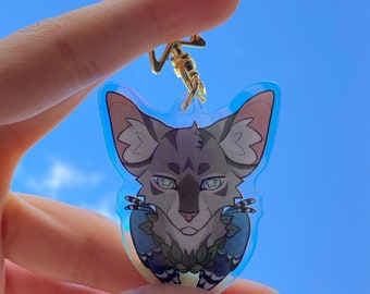 May include: A clear acrylic keychain featuring a cartoon illustration of a gray and white cat with blue eyes. The cat is wearing a blue and white patterned outfit with green leaves around its neck. The keychain has a gold metal ring and a gold metal chain.