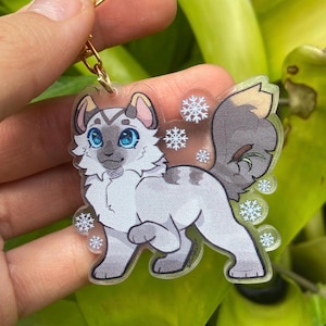 Puede incluir: Un llavero de acrílico transparente con un gato de dibujos animados de pelaje blanco, marcas grises y ojos azules. El gato camina y está rodeado de copos de nieve.
