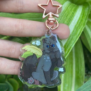 Op de afbeelding: Een heldere acryl sleutelhanger met een cartoonkat met blauwe ogen, zittend bij een groen blad en witte bloemen. De sleutelhanger heeft een roségouden stervormige sluiting.