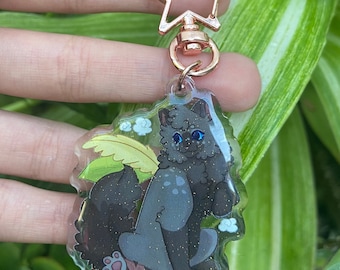 May include: A clear acrylic keychain featuring a cartoon cat with blue eyes, sitting near a green leaf and white flowers. The keychain has a rose gold star-shaped clasp.