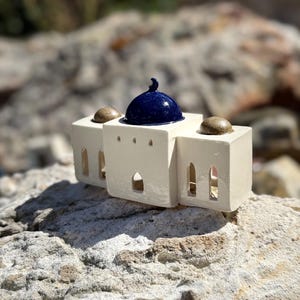 Peut inclure: Une miniature de mosquée en céramique, de couleur crème, avec un dôme bleu foncé. Elle présente des fenêtres cintrées et deux petits dômes arrondis. La pièce est posée sur une surface rocheuse texturée.