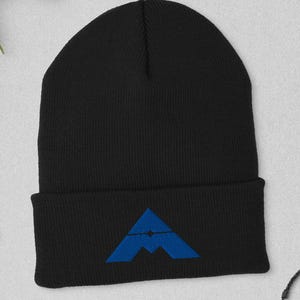 May include: Black beanie with a blue mountain-shaped logo. The beanie is made of a soft material and has a folded cuff. The hat is displayed on a light gray surface with a watch, glasses, and a wallet.