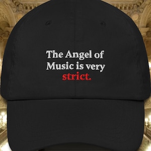 Może przedstawiać: Czarna czapka bejsbolówka z haftowanym napisem "The Angel of Music is very strict." w kolorze białym i czerwonym. Czapka ma zakrzywiony daszek i strukturalny krój, odpowiednia do noszenia na co dzień.