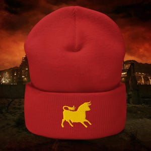 Pode incluir: Um gorro vermelho com um logotipo de touro bordado em amarelo na aba dobrada. O gorro é feito de um material de malha macia. O fundo mostra uma paisagem escura e dramática.