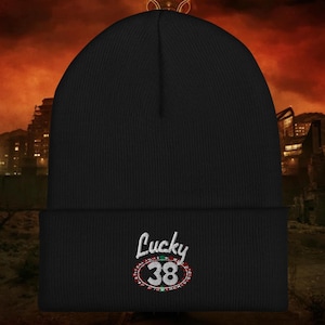 May include: Black beanie with the text "Lucky 38" embroidered in white script and a red, green, and white circular design. The beanie is made of a ribbed knit material. The background shows a city skyline with a red and orange sky.