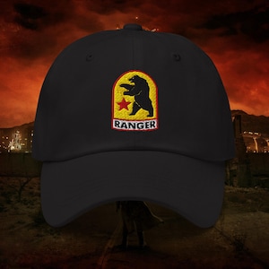 May include: Black baseball cap with a patch featuring a bear, a red star, and the word "RANGER" in white lettering. The patch is set against a yellow background. The cap has a curved brim and a button on top.