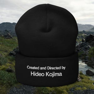 Pode incluir: Gorro preto com o texto "Created and Directed by Hideo Kojima" bordado em branco. O gorro é um chapéu de malha macia, perfeito para o clima frio. O chapéu é um acessório elegante.