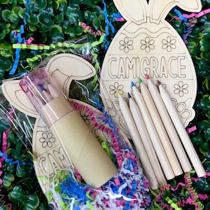 May include: A wooden Easter egg and bunny-shaped craft kit with the name "CAMIGRACE" engraved on the egg. The kit includes coloured pencils and a roll of craft supplies, all packaged with colourful confetti and set against a green backdrop.