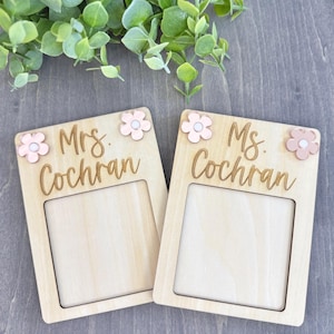 Puede incluir: Dos placas de madera con los textos "Mrs. Cochran" y "Ms. Cochran" en cursiva. Cada placa tiene una abertura rectangular y está decorada con detalles de flores rosas. El fondo es una superficie de madera gris.