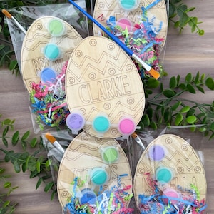 May include: Wooden Easter egg paint kits with paint pots, paintbrushes, and colourful paper shred. Each egg is engraved with a name. The eggs are packaged in clear cellophane bags. The names on the eggs are Clarke, Tom, RDY, and Isabella.