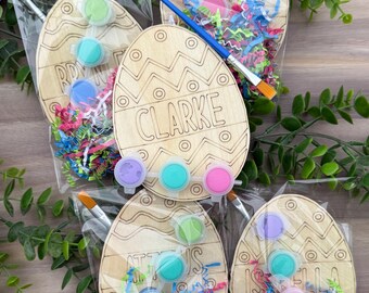 May include: Wooden Easter egg paint kits with paint pots, paintbrushes, and colorful paper shred. Each egg is engraved with a name. The eggs are packaged in clear cellophane bags. The names on the eggs are Clarke, Tom, RDY, and Isabella.