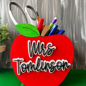 Puede incluir: Organizador de escritorio en forma de manzana roja con una hoja verde y el nombre "Mrs. Tomlinson" en letras blancas y negras. El organizador está lleno de bolígrafos, marcadores y tijeras.