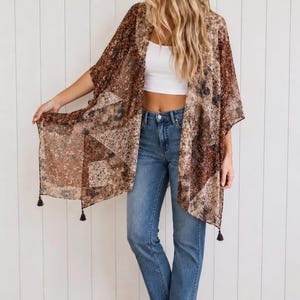 May include: A sheer, open-front kimono featuring a patchwork design of various floral patterns in shades of brown, tan, and cream. The garment has wide, loose sleeves and is finished with small, dark brown tassels along the bottom hemline.