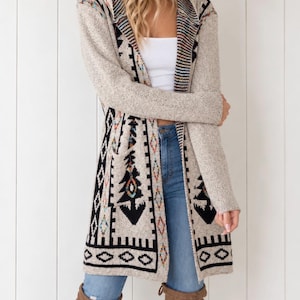 May include: A long-sleeved, open-front cardigan in a heathered beige knit. The garment features a prominent black, white, and multi-colored geometric pattern along the lapels, hem, and pockets, with a repeating arrow and diamond motif.