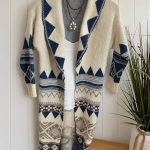 May include: A long, open-front knit cardigan featuring a cream base with navy blue and brown geometric patterns. The garment has a wide shawl collar and long sleeves, layered over a plain white tank top and a multi-strand beaded necklace.