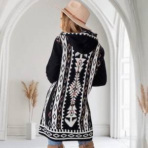 May include: A black hooded cardigan with a geometric pattern in white, red, yellow, and blue. The long sweater features a detailed design on the back and a hood. The model is wearing a tan hat.