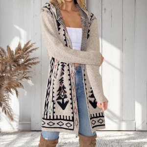 May include: A long, open-front cardigan in a neutral beige color with a patterned design in black, teal, and red. The cardigan features a hood and pockets. The design includes geometric and tribal-inspired motifs. The sleeves are solid beige.