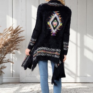 May include: Black open-front cardigan with a colorful geometric design on the back and trim. The long-sleeved sweater has a soft, fuzzy texture and a flowing, asymmetrical hemline. The design includes white, yellow, red, and blue accents.
