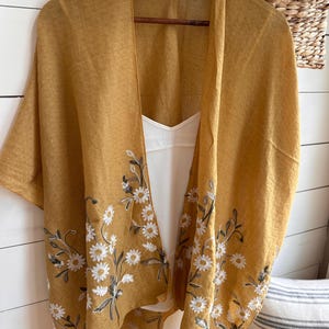 May include: Mustard yellow kimono-style cover-up with white daisy floral embroidery. The lightweight garment is hanging on a wooden hanger, revealing a white camisole underneath. A straw hat and woven basket are visible in the background.