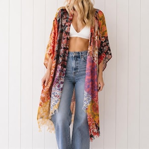 May include: A long, open-front kimono featuring a vibrant patchwork of floral and paisley patterns in shades of orange, yellow, pink, black, and white. The garment has wide, loose sleeves and a frayed hemline, worn over a white bralette and blue jeans.
