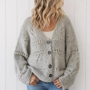 May include: A light gray, chunky knit cardigan featuring a V-neckline, long sleeves, and four dark buttons down the front. The garment displays a textured pattern with open-knit sections across the chest and sleeves, paired with blue denim jeans.