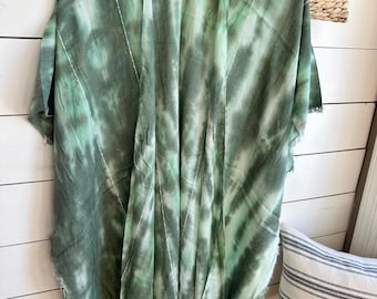 Pode incluir: Um kimono verde tie-dye com bainha com franjas está pendurado em um cabide de madeira. O tecido apresenta tons de verde e branco, com detalhes de costura brancos verticais. A peça de roupa é exibida contra uma parede de painéis de madeira branca.