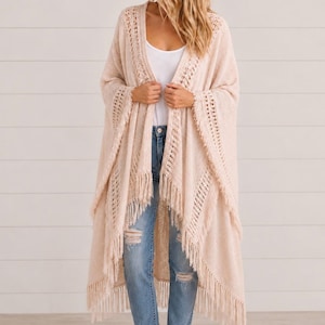 May include: A beige open-front cardigan featuring an intricate crochet pattern along the lapels and sleeves, finished with long fringe trim along the hemline. The garment is worn over a white tank top and distressed blue denim jeans.