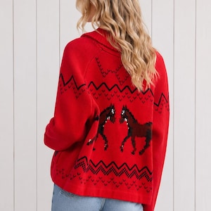 May include: A bright red long-sleeve knit sweater featuring a black geometric zigzag pattern across the upper and lower sections, with two dark brown horse figures centered on the back. The sweater has a fold-over collar and a loose fit.