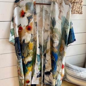 May include: A floral print kimono in shades of white, blue, yellow, and red. The lightweight, open-front garment is hanging on a wooden hanger. The design features an abstract watercolor floral pattern.
