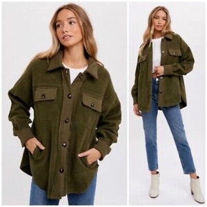 May include: An olive green button-down jacket with two chest pockets and two side pockets. The jacket has a collared neckline and long sleeves. The jacket is paired with blue jeans. The jacket is made of a soft, textured fabric.