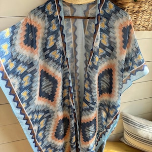 May include: A lightweight, open-front kimono with a geometric pattern in shades of blue, orange, and yellow. The kimono is hanging on a wooden hanger, with a straw hat visible above. The design features a zigzag border.