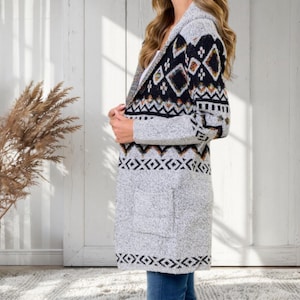 May include: A long, gray cardigan with a hood and pockets. The upper portion features a black, white, and orange geometric pattern. The lower portion has a repeating geometric design. The cardigan is worn over a white top and blue jeans.