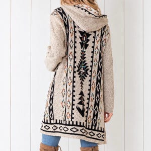 May include: A beige hooded cardigan featuring a black, orange, and blue geometric pattern on the back. The knit garment is worn with a beige wide-brimmed hat, blue denim jeans, and brown suede over-the-knee boots with lace-up details.