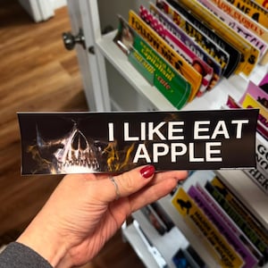 May include: A black bumper sticker with the text "I LIKE EAT APPLE" in white, featuring a skull graphic with yellow flames. The sticker is held in a hand with a red painted nail.