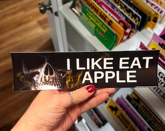 Może przedstawiać: Czarna naklejka na zderzak z białym napisem "I LIKE EAT APPLE", z grafiką czaszki z żółtymi płomieniami. Naklejka jest trzymana w dłoni z pomalowanym na czerwono paznokciem.