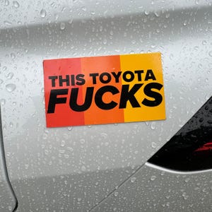 Könnte beinhalten: Ein rechteckiger Aufkleber mit dem Text "THIS TOYOTA FUCKS" in Schwarz, vor einem Farbverlauf aus Rot, Orange und Gelb. Der Aufkleber ist auf einem silbernen Auto angebracht, Wassertropfen sind sichtbar.