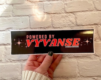 May include: A black sticker with the text "POWERED BY VYVANSE" in pink and red, with starburst accents. The sticker is held by a person in front of a white brick wall.