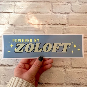 May include: A rectangular sticker with the text "POWERED BY ZOLOFT" in a retro font, with yellow starburst graphics on a light blue background. The sticker is held against a white brick wall.