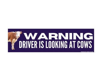 May include: A rectangular sticker with a dark blue background and white text that reads "WARNING DRIVER IS LOOKING AT COWS." A brown and white cow is on the left side of the sticker.