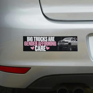 May include: A rectangular bumper sticker with a black background featuring the white and pink text BIG TRUCKS ARE GENDER AFFIRMING CARE with two pink hearts and a white pickup truck on the right side, applied to a silver vehicle bumper.